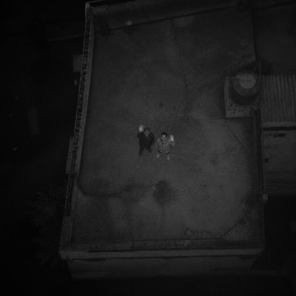 Overhead low-light drone view of two survivors waving from a rooftop